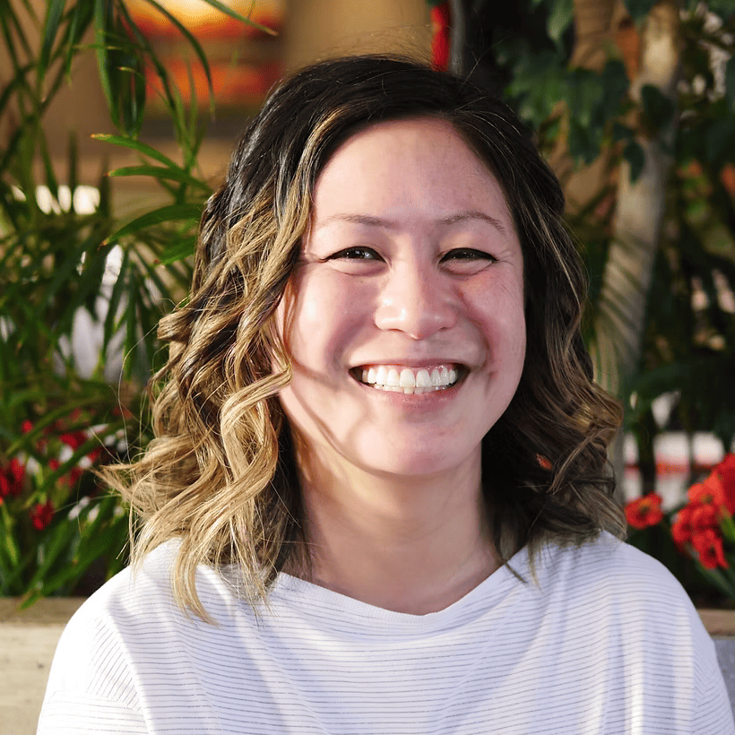 Smiling woman with wavy hair in white top standing in front of greenery