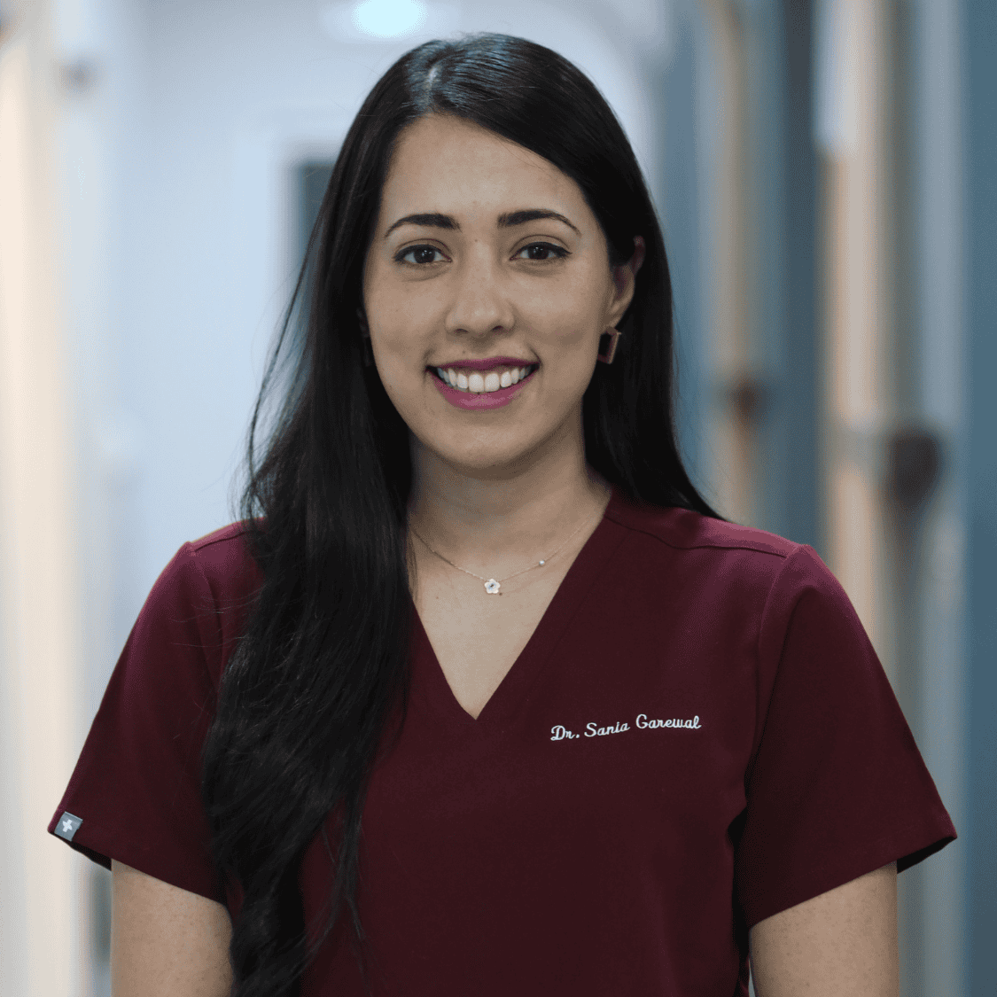 Dr. Sonia Garewal smiling in a burgundy scrub top, standing in a dental clinic hallway